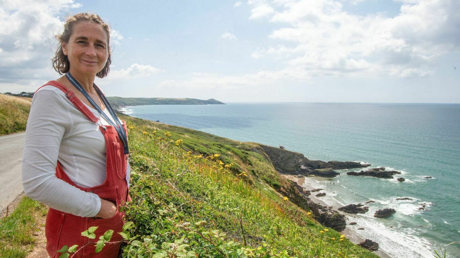 Debt Centre Manager Melissa, a white lady with shoulder length brown hair, in red dungarees and a grey long sleeved top. She is smiling at the camera, hands in pockets, overlooking the beautiful Cornish coast.