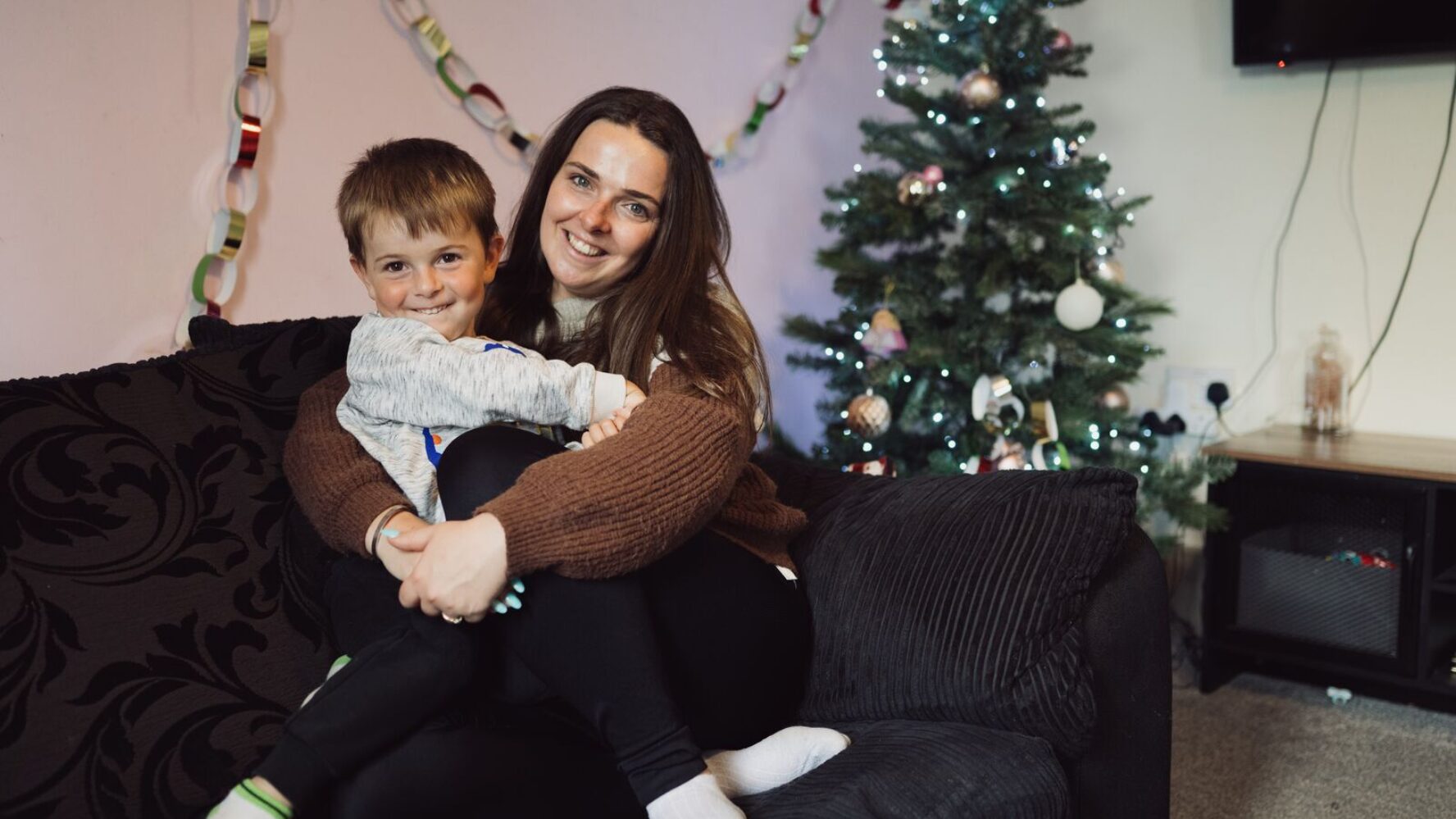 CAP client, Jade, sat on her sofa cuddling her son and smiling, with Christmas decorations and a Christmas tree behind them .