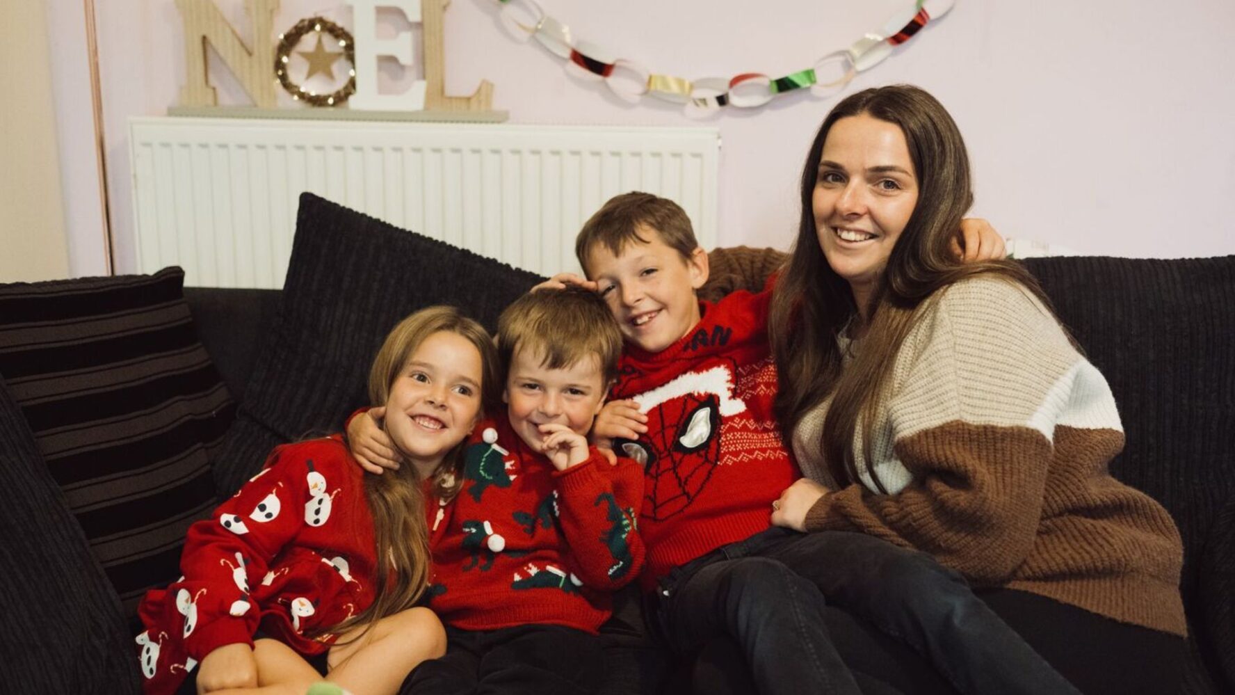 Jade with her three children, Layla, Lukas and Jacob. They're sat on the sofa all cuddling together in Christmas jumpers, with decorations up behind them. They're all smiling at the camera.